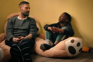 Ein Kind und ein Mann sitzen auf einem Riesenkuscheltier und schauen sich schweigend an