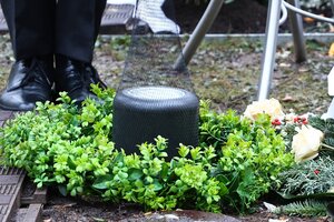 Eine Urne wird in die Erde gelassen zwischen einem grünen Kranz, schwarze Schuhe sind zu sehen