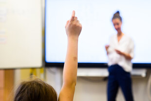 Ein Schüler meldet sich per Handzeichen während eine Lehrerin vor einer digitalen Schultafel im Klassenraum einer 4. Klasse einer Grundschule steht