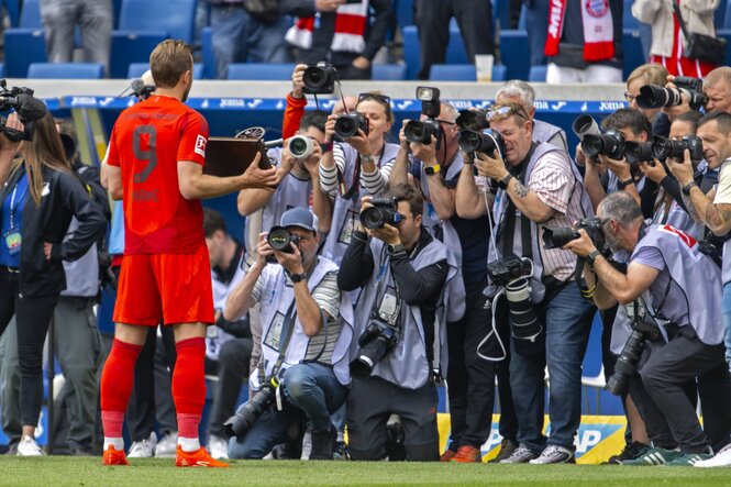 Zu sehen ist ein Fussballspieler auf dem Feld vor einem Haufen Journalisten, die ihn fotografieren.