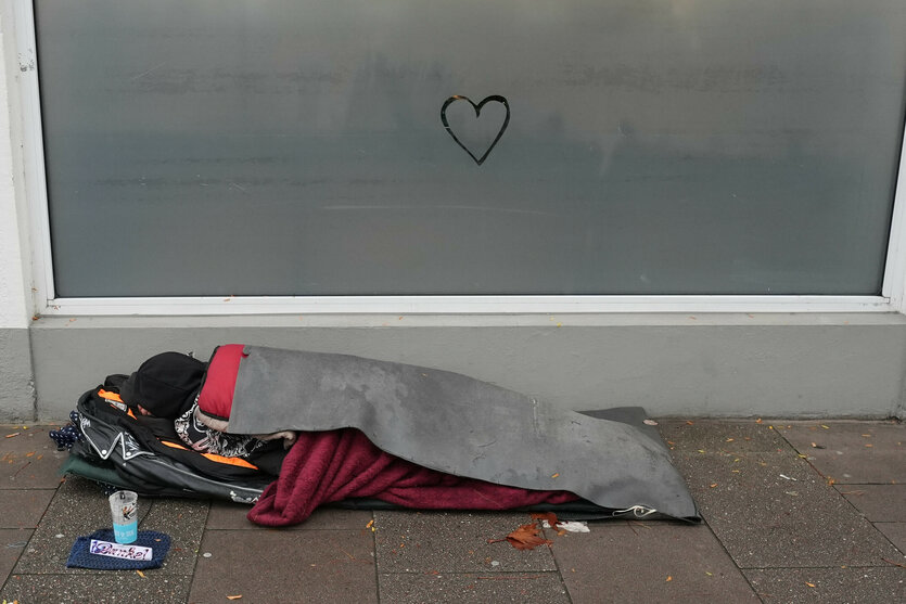 Ein junger Mensch liegt unter einer grauen Decke auf dem Gehweg