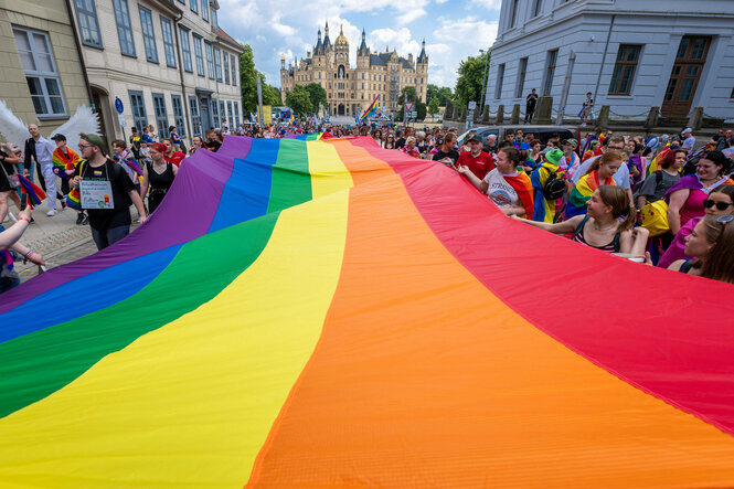 Ein großer Regenbogenfahne wird von vielen Menschen bei einem Umzug durch eine Stadtstraße getragen. Im Hintergrund steht ein prächtiges, schlossartiges Gebäude mit Türmen und goldenen Kuppeln. Die Menge ist bunt gekleidet, einige tragen Regenbogenaccessoires, und die Atmosphäre wirkt fröhlich und feierlich.
