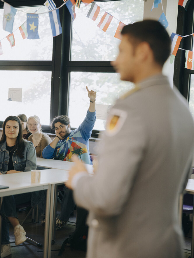 Julian Ströbl steht in Uniform vor einer Schulklasse, ein Schüler meldet sich