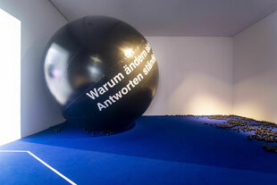 Ein großer, schwarzer Ball mit der weißen Aufschrift „Warum ändern sich Antworten ständig?“ liegt in einem Raum mit weißen Wänden und blauem Boden. Viele kleine schwarze Kugeln sind auf dem Boden verstreut. Licht fällt durch ein Fenster links und erzeugt