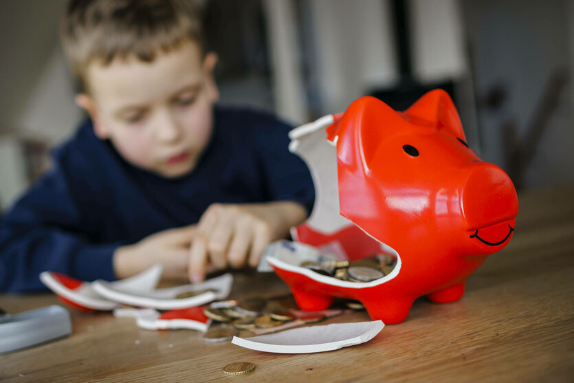 Ein Junge hinter einer zerschlagemem Sparschein, aus welchem er das enthaltene Geld zählt