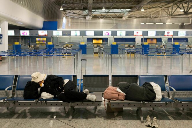 Zwei Passagiere schlafen auf einer FLughafenbank, der Flughafen scheint ausgestorben und leer.
