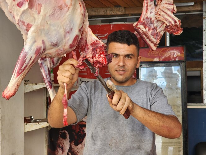 Adham in seiner Fleischerei, im Hintergrund ein Kühlschrank, er selbst schneidet etwas von einem herabhängenden Stück Fleisch ab