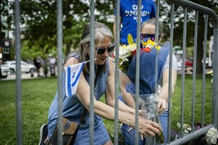 Zwei Frauen befestigen Blumen an einem Zaun in der Nähe des Tagtorts, eine der Frauen halt eine kleine israelische Flagge mit den Zähnen fest
