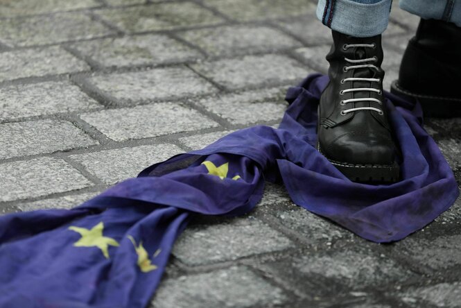 Eine Europaflagge liegt auf der Straße, ein Mann hat seinen Stiefel mit weissen Schnürsenkeln draufgestellt