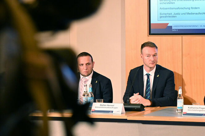 Zwei Personen sitzen an einem Tisch bei einer Konferenz, vor ihnen Mikrofone und Namensschilder. Im Hintergrund ist ein Bildschirm mit Text über Antisemitismusbekämpfung und Gedenkkultur. Eine Kamera ist im Vordergrund sichtbar.