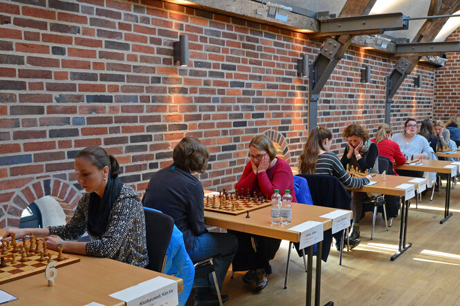 An einer Backsteinwand stehen sechs Tische, an denen Frauen gegeneinander Schach spielen