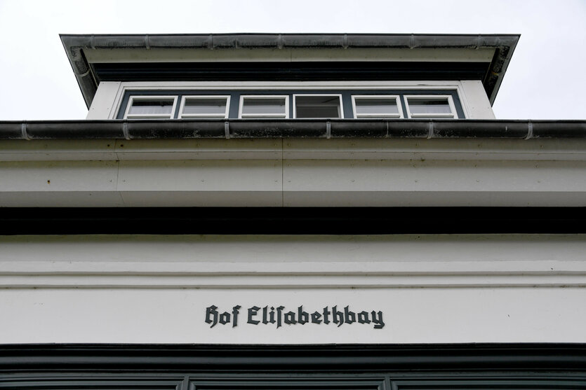 Ein weißes Gebäude mit schwarzem Dach und einer Inschrift "Hof ELISABETHBAY" in gotischer Schrift. Oben ein Fenster mit mehreren Scheiben. Der Himmel ist hell und klar.