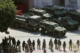 Hinter uniformierten bewaffneten Männern rollen Gepanzerte Fahrzeuge eine Straße entlang