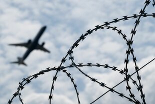 Blick auf ein startendes Flugzeug, im Vordergrund ist Stacheldraht zu sehen