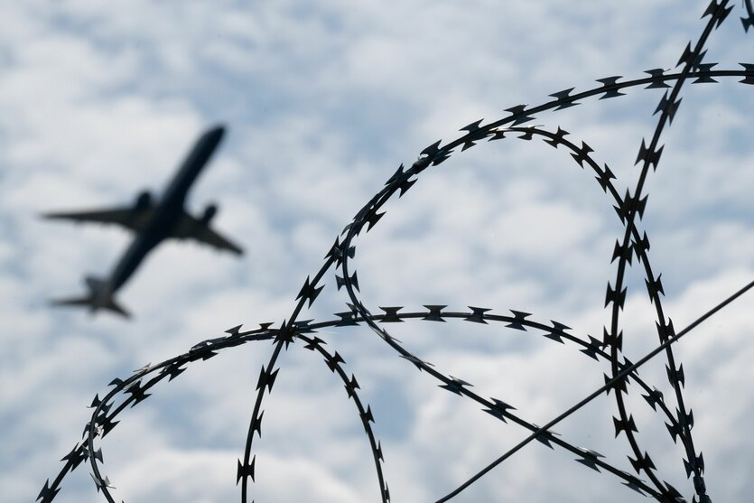 Blick auf ein startendes Flugzeug, im Vordergrund ist Stacheldraht zu sehen