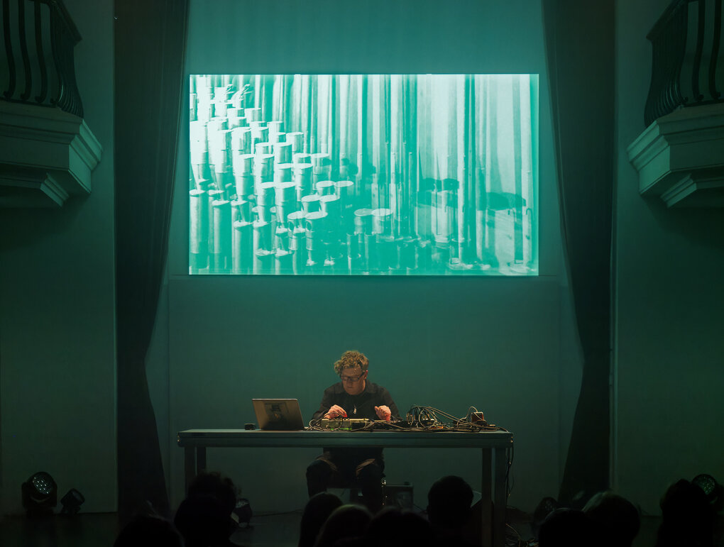 Der Musiker Hanno Leichtmann sitz an einem Tisch, vor ihm eine Computer und diverese Mischpulte und Kabel. Im Hintergrund ist das Cover von "Outerlands" in grünes Licht getaucht auf die Wand projiziert.