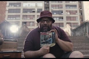 Ein Mann mit Cappy sitzt auf einer Treppe vor einem Hochhauskomplex. Er hält stolz eine Vinylsingle von Darko Domijan in die Kamera.