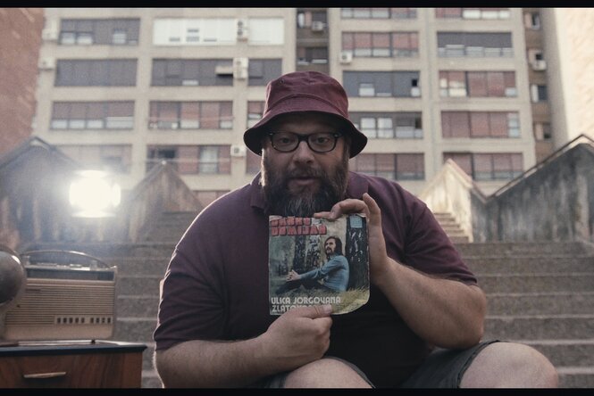 Ein Mann mit Cappy sitzt auf einer Treppe vor einem Hochhauskomplex. Er hält stolz eine Vinylsingle von Darko Domijan in die Kamera.