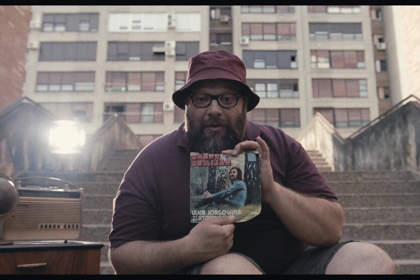 Ein Mann mit Cappy sitzt auf einer Treppe vor einem Hochhauskomplex. Er hält stolz eine Vinylsingle von Darko Domijan in die Kamera.