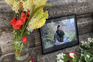 Ein Foto zeigt einen Blumenstrauß mit gelben und roten Blüten in einer Vase, neben einem gerahmten Bild einer sitzenden Person im Freien, umgeben von weiteren Blumen und einer Kerze auf steinernem Untergrund.