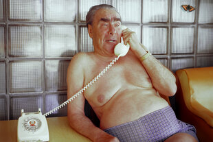 Ein Mann sitzt nur mit einer Unterhose bekleidet auf einem Stuhl und telefoniert mit einem alten Wählscheibentelefon