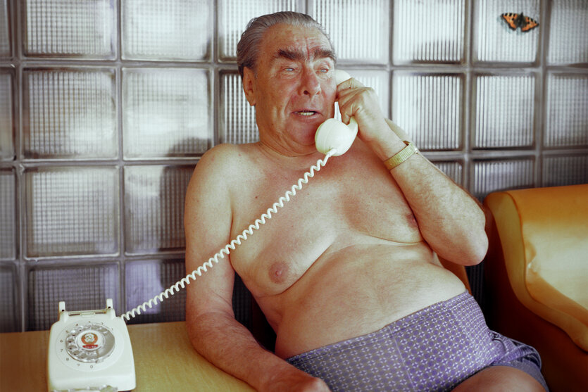 Ein Mann sitzt nur mit einer Unterhose bekleidet auf einem Stuhl und telefoniert mit einem alten Wählscheibentelefon