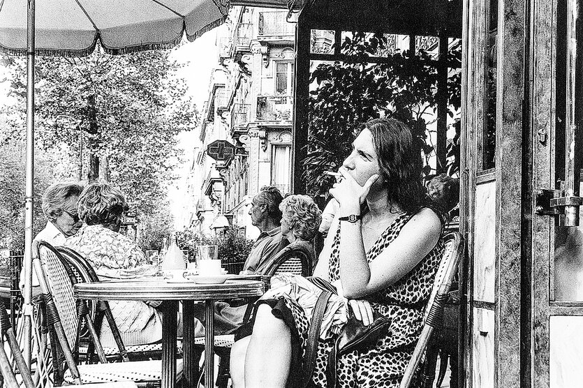 Eine junge Frau im ärmellosen Kleid sitzt rauchend in einem Straßencafe