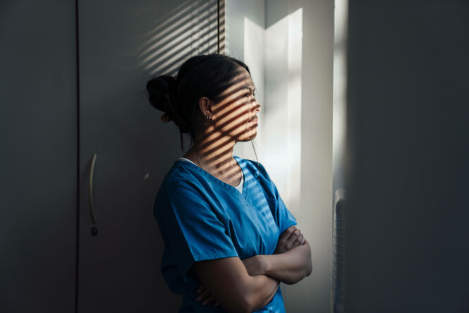 Krankenschwester in blauer Uniform macht eine Pause und schaut aus dem Fenster, während das Sonnenlicht Schatten in den Raum wirft