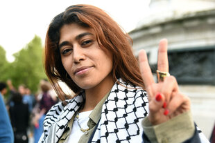 Porträt von Rima Hassan, sie schaut kämpferisch in die Kamera, macht das Peacezeichen und trägt Kuffiah