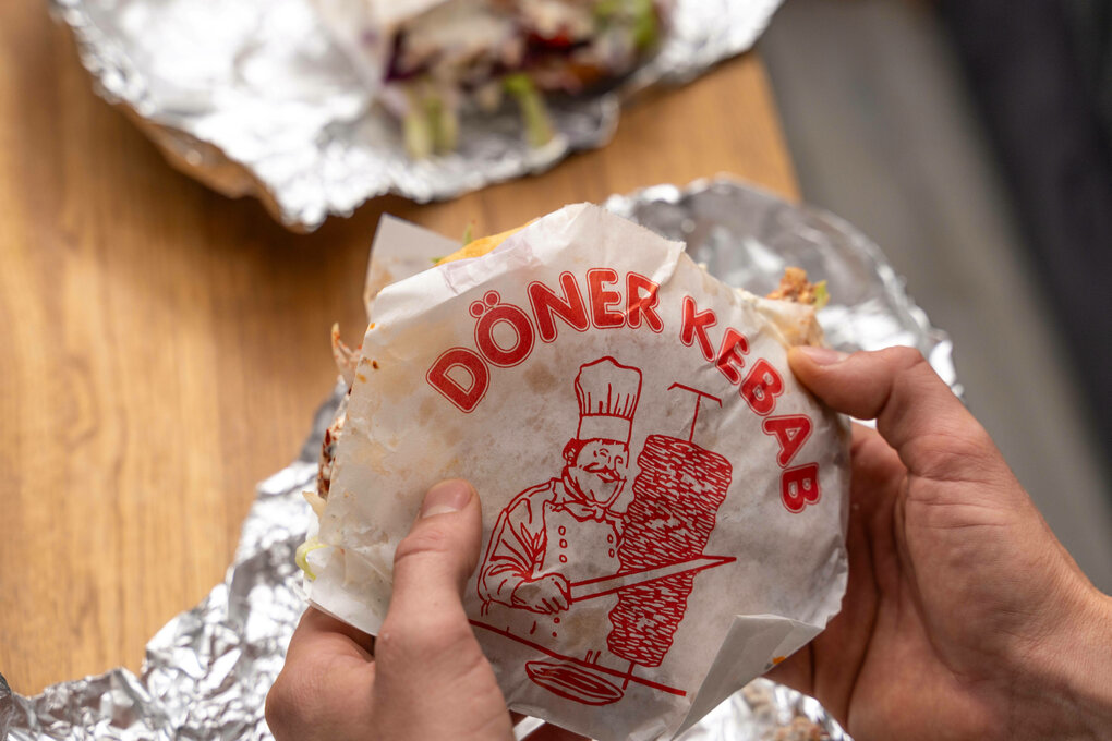 Ein Mann hält einen vegetarischen Döner Kebap ohne Fleisch in der Hand. Auf dem Esstisch Aluminium Folie von der Verpack