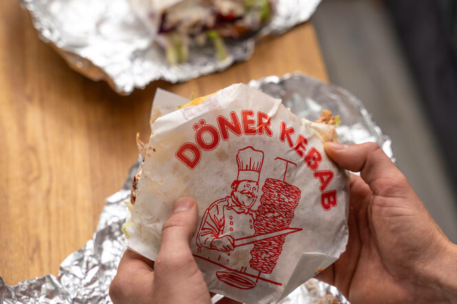 Ein Mann hält einen vegetarischen Döner Kebap ohne Fleisch in der Hand. Auf dem Esstisch Aluminium Folie von der Verpack