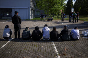 Rückansicht von Menschen, die auf einem Parkplatz in der Reihe teilweise hocken oder sitzen. Im Hintergrund ist ein Polizist erkennen, der sich mit anderen Menschen unterhält