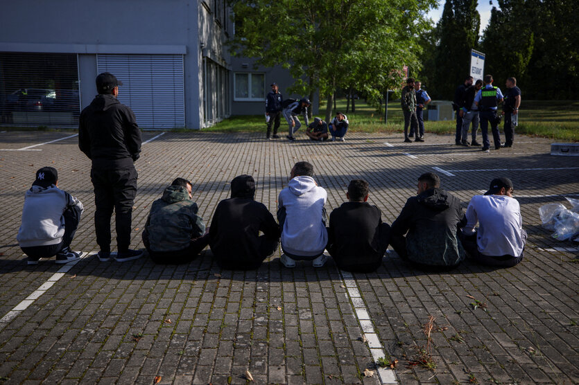 Rückansicht von Menschen, die auf einem Parkplatz in der Reihe teilweise hocken oder sitzen. Im Hintergrund ist ein Polizist erkennen, der sich mit anderen Menschen unterhält