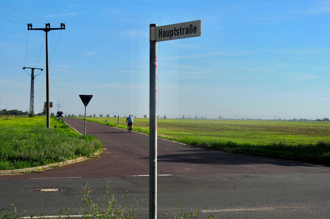 Einsame Straßenkreuzung unter blauem Himmel, im Hntergrund entfernt sich ein Mann auf einem Fahrrad, im Vordergrund ein Schild: "Hauptstraße"