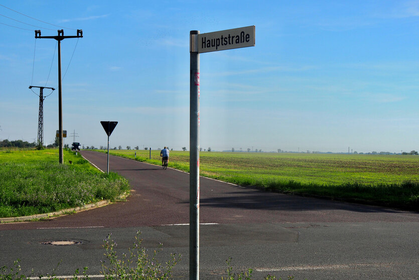 Einsame Straßenkreuzung unter blauem Himmel, im Hntergrund entfernt sich ein Mann auf einem Fahrrad, im Vordergrund ein Schild: "Hauptstraße"