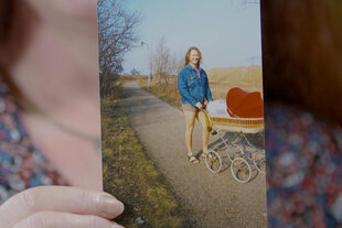 Eine Frau hält ein Foto, darauf ein Mann mit kurzen Hosen und langen Haaren, der einen Kinderwagen schiebt