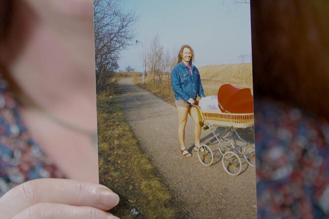Eine Frau hält ein Foto, darauf ein Mann mit kurzen Hosen und langen Haaren, der einen Kinderwagen schiebt