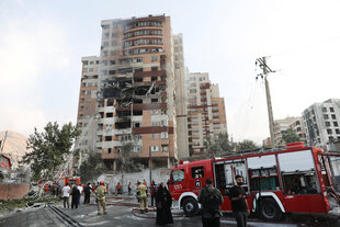 Ein Wohnhochhaus mit schweren Schäden an der Fassade. Auf der Straße stehen Feuerwehrfahrzeuge, drumherum Menschen, eine Feuerwehrleiter ragt in die Höhe
