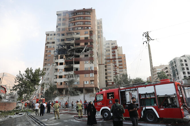 Ein Wohnhochhaus mit schweren Schäden an der Fassade. Auf der Straße stehen Feuerwehrfahrzeuge, drumherum Menschen, eine Feuerwehrleiter ragt in die Höhe