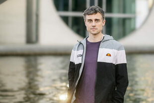Ein junger Mann mit kurzer, grau melierter Frisur steht in sportlicher Jacke am Wasser vor moderner Architektur, dem Bundestag in Berlin. Nick Jaussi blickt ruhig und direkt in die Kamera.