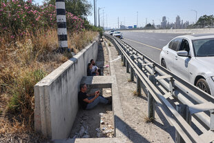 Hinter der Leitplanke einer Straße, angelehnt an eine Mauer, die die Straße begrenzt, sitzen Menschen