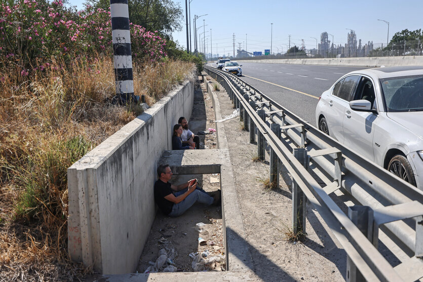 Hinter der Leitplanke einer Straße, angelehnt an eine Mauer, die die Straße begrenzt, sitzen Menschen
