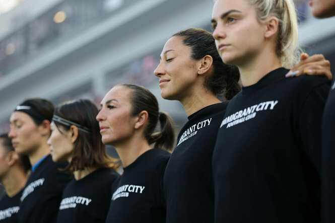 Spielerinnen Arm in Arm mit einem T-Shirt mit dem Aufdruck "Immigrant City Football Club"