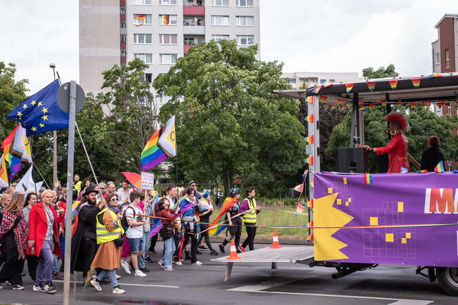 Menschen in bunter Kleidung laufen hinter einem Demo-Truck, im Hintergrund ein Hochhaus
