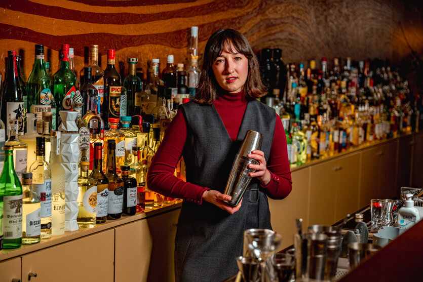 Eine Frau mit mittellangem braunem Haar steht in ihrer Bar, im Hintergrund zahllose Spirituosen auf einem langen Schrank