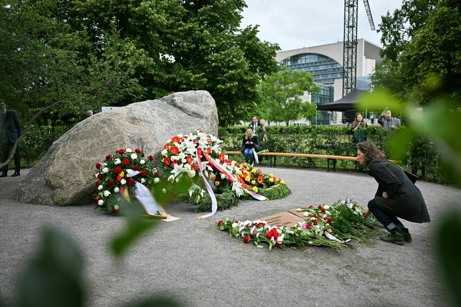 Eine Person steht in der Hocke vor einem Gedenkstein und Kränzen mit überwiegend rot-weißen Blumen