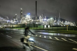 Ein Radfahrer fährt im Dunkeln eine Straße entlang, im Hintergrund die Lichter einer Fabrik