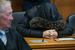 Ein Mann, der sich die pelzbesetzte Kapuze eines Anoraks über das Gesicht gezogen hat, sitzt auf einer Anklagebank