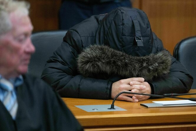 Ein Mann, der sich die pelzbesetzte Kapuze eines Anoraks über das Gesicht gezogen hat, sitzt auf einer Anklagebank