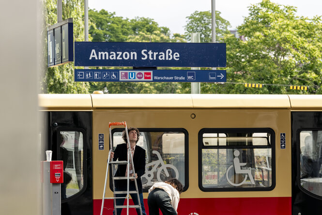 An einem S-Bahnhof sind die Stationsnamensschilder mit der Aufschrift „Amazon Straße“ überklebt, darunter der Künstler mit einer Leiter